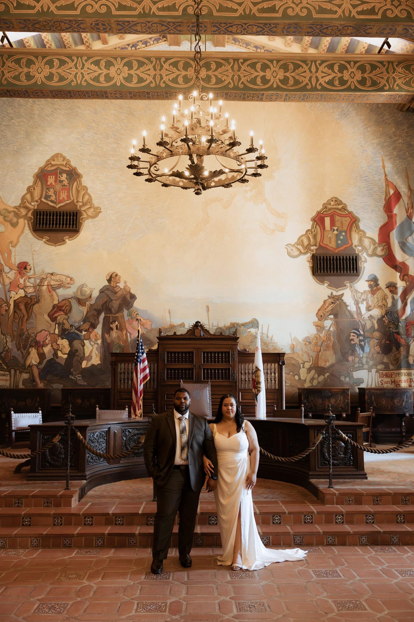 Couple under the chandelier and 1920s murals of the Santa Barbara courthouse Mural Room